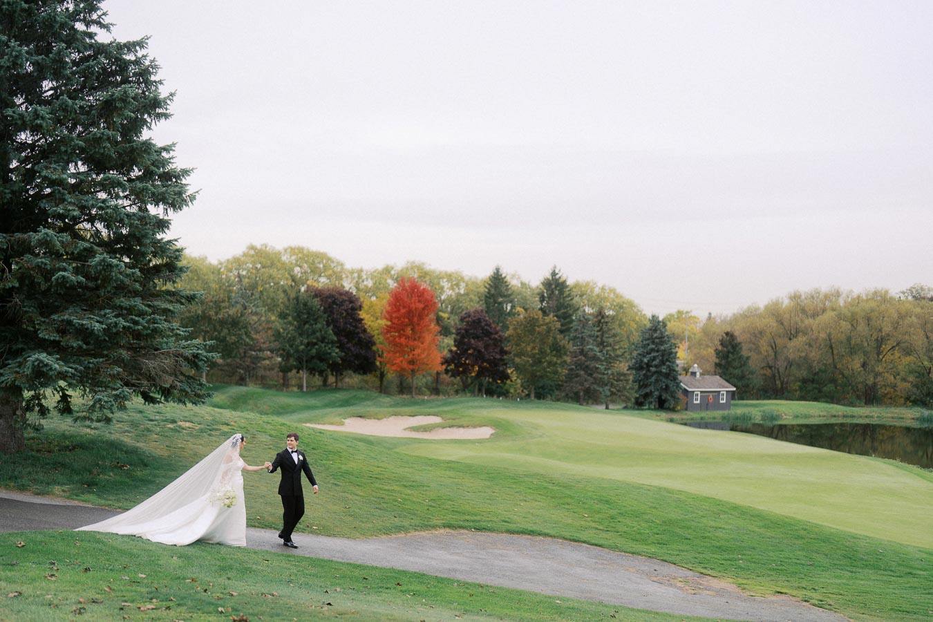 Bride and groom walking hand in hand on a golf course with autumn trees in the background, showcasing a picturesque outdoor wedding setting.