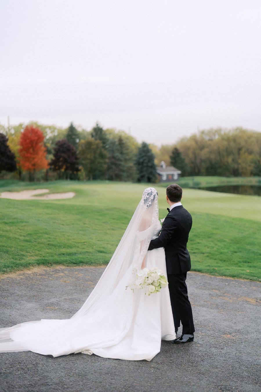 Bride and groom in elegant attire standing together on a golf course, amidst lush greenery and autumn-colored trees, capturing a serene outdoor wedding moment.