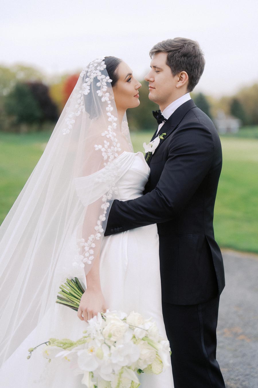 Bride and groom embracing on their wedding day, with the bride in an elegant white gown holding a bouquet of white flowers, and the groom in a classic black tuxedo, against a scenic outdoor background.
