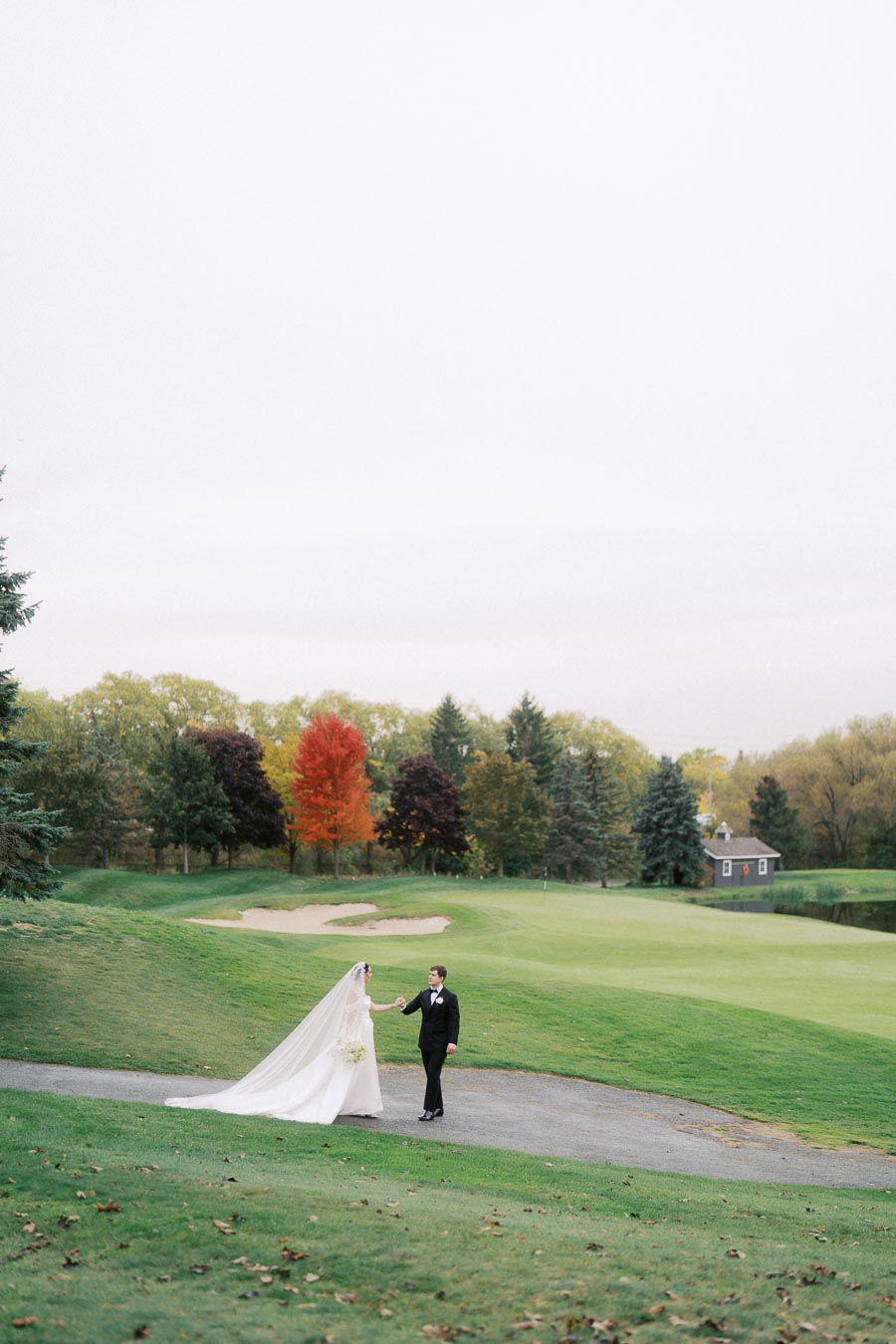 Bride and groom walking on a path in a scenic outdoor setting, surrounded by lush green grass and autumn trees, with a rustic building in the background.