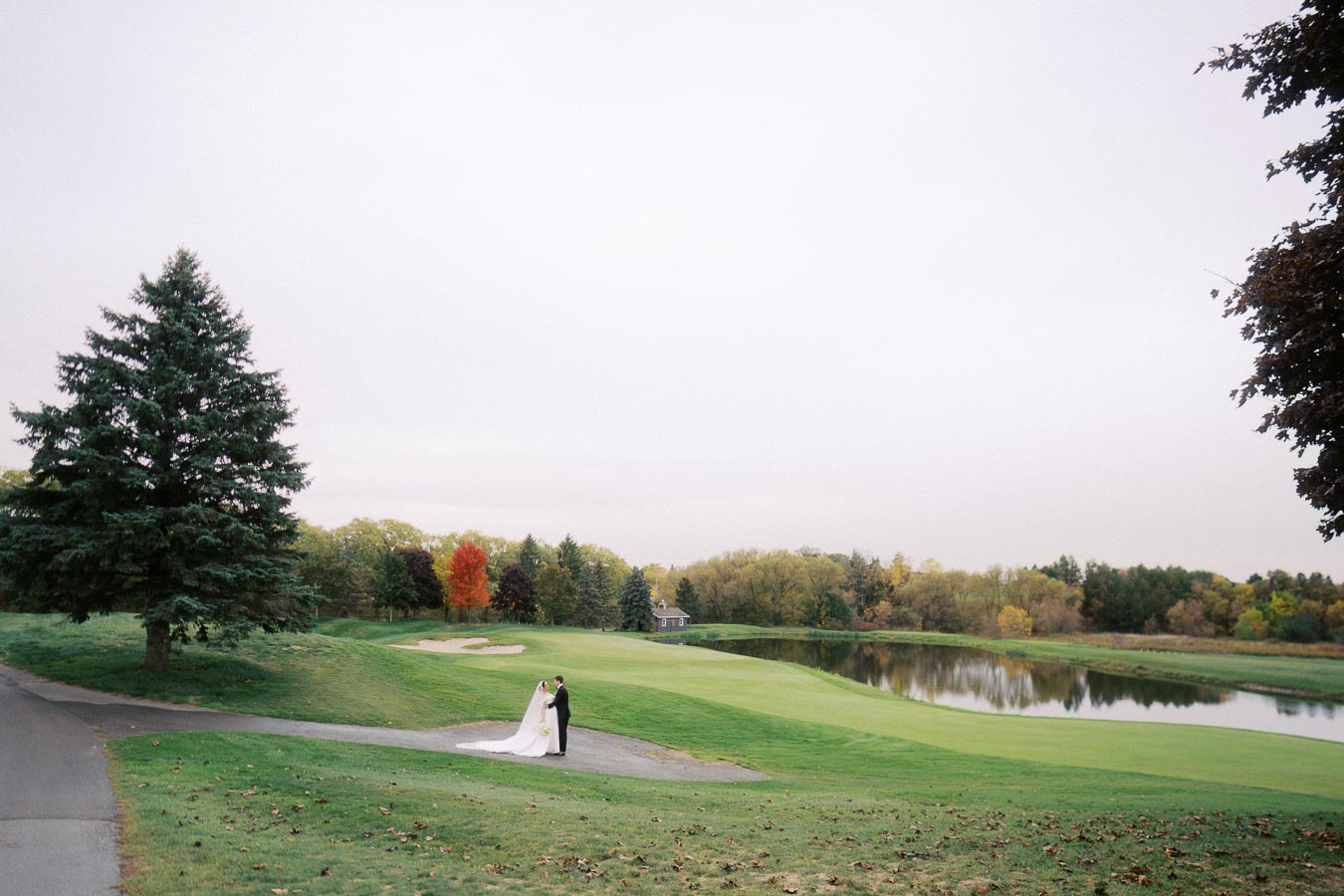 Bride and groom embracing on a scenic golf course with lush greenery, a serene pond, and autumn trees in the background, creating a picturesque wedding setting.