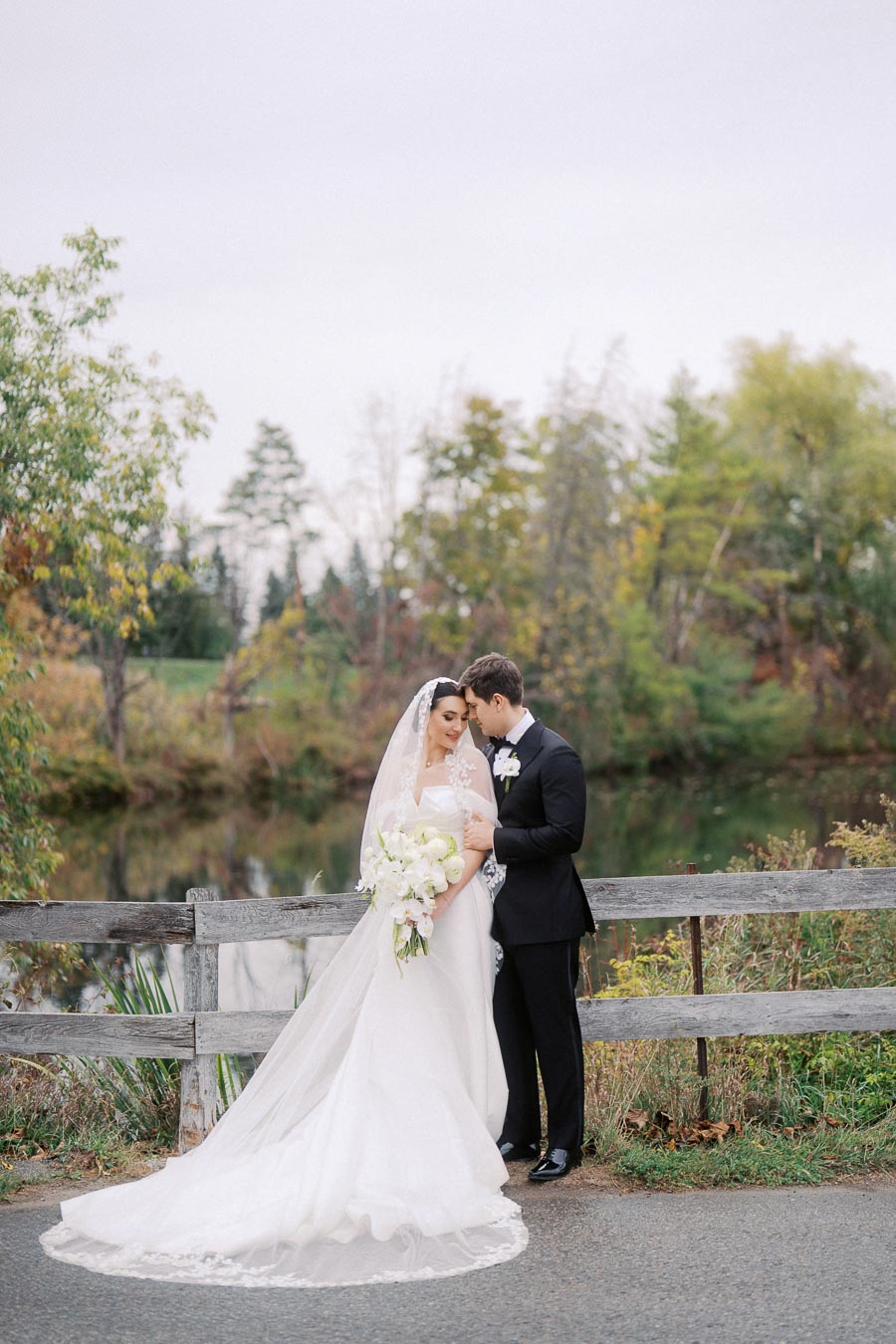A bride in a white wedding dress and veil holds a bouquet while standing next to a groom in a black suit, set against a picturesque outdoor backdrop with trees and a serene pond.