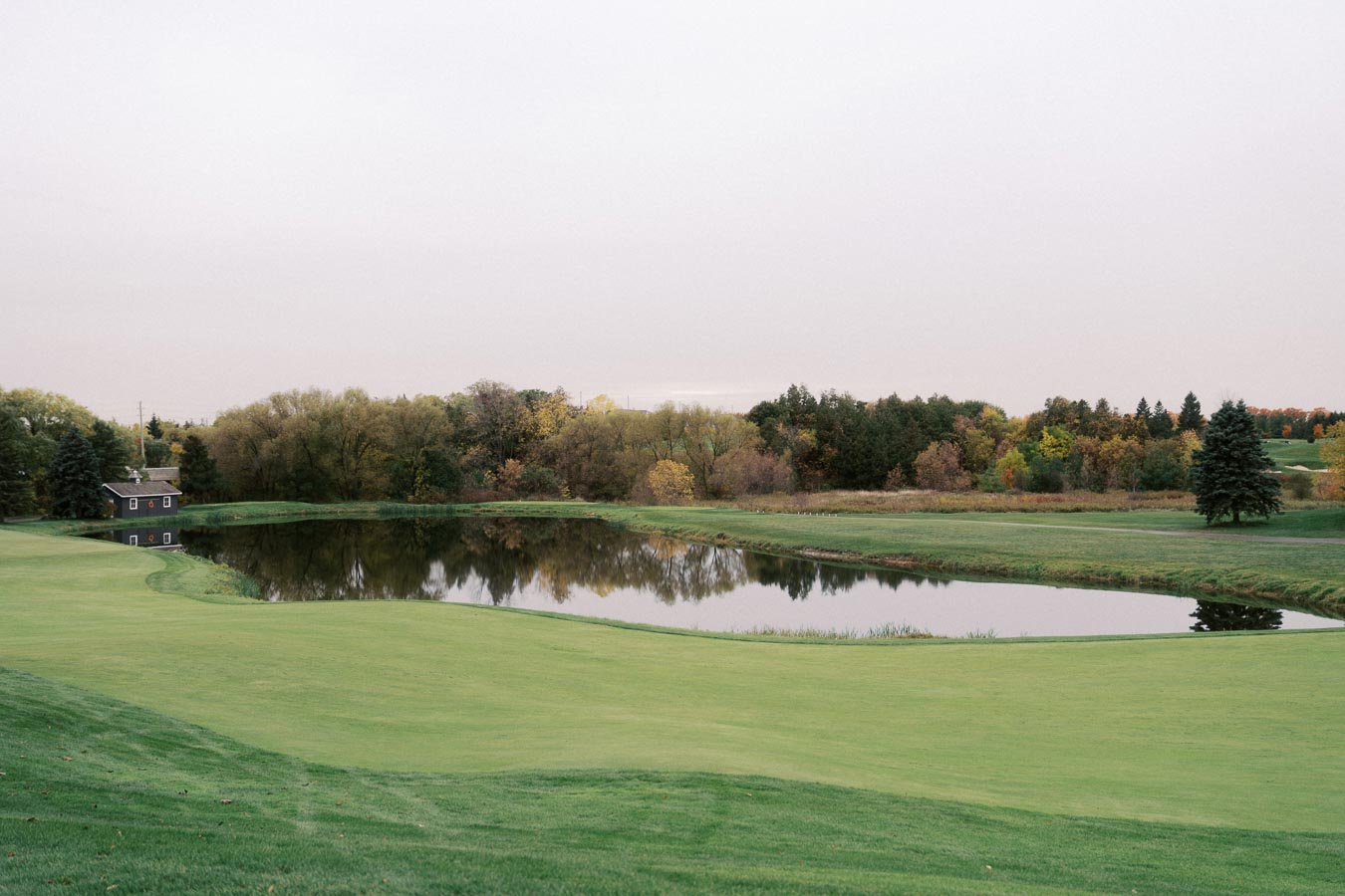A scenic view of a lush green golf course featuring a serene pond surrounded by autumn-colored trees under a clear sky.