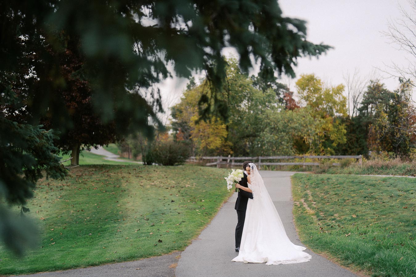Bride and groom embrace on a scenic outdoor path surrounded by lush greenery and trees, with the bride in a white gown holding a bouquet of white flowers.