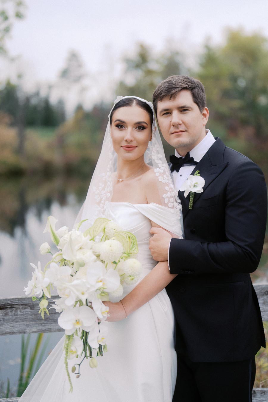 Bride and groom in elegant wedding attire, posing outdoors by a serene pond, with the bride holding a bouquet of white flowers and wearing a detailed veil.