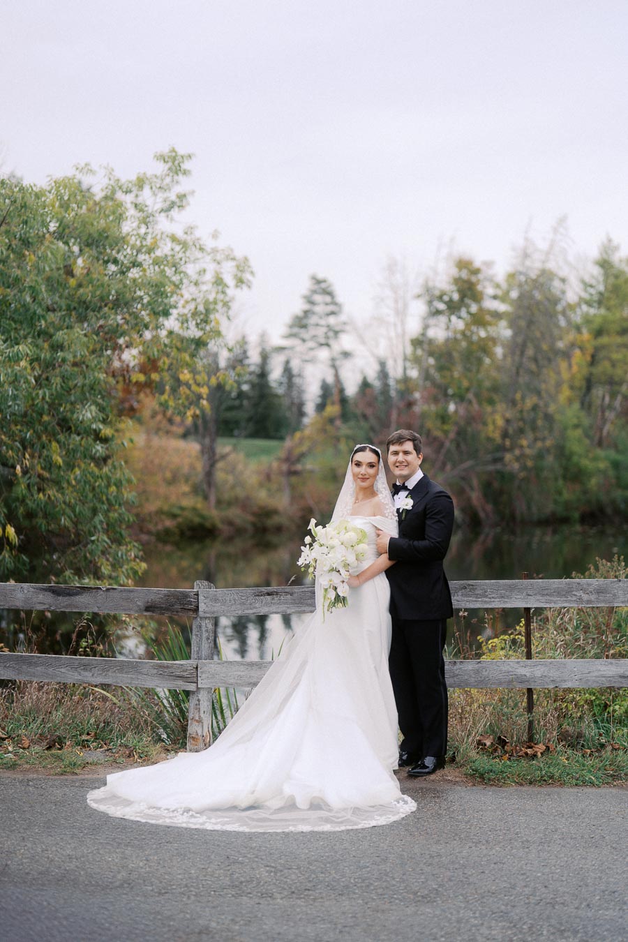 Bride and groom pose in elegant wedding attire by a picturesque pond with lush greenery and rustic wooden fence.