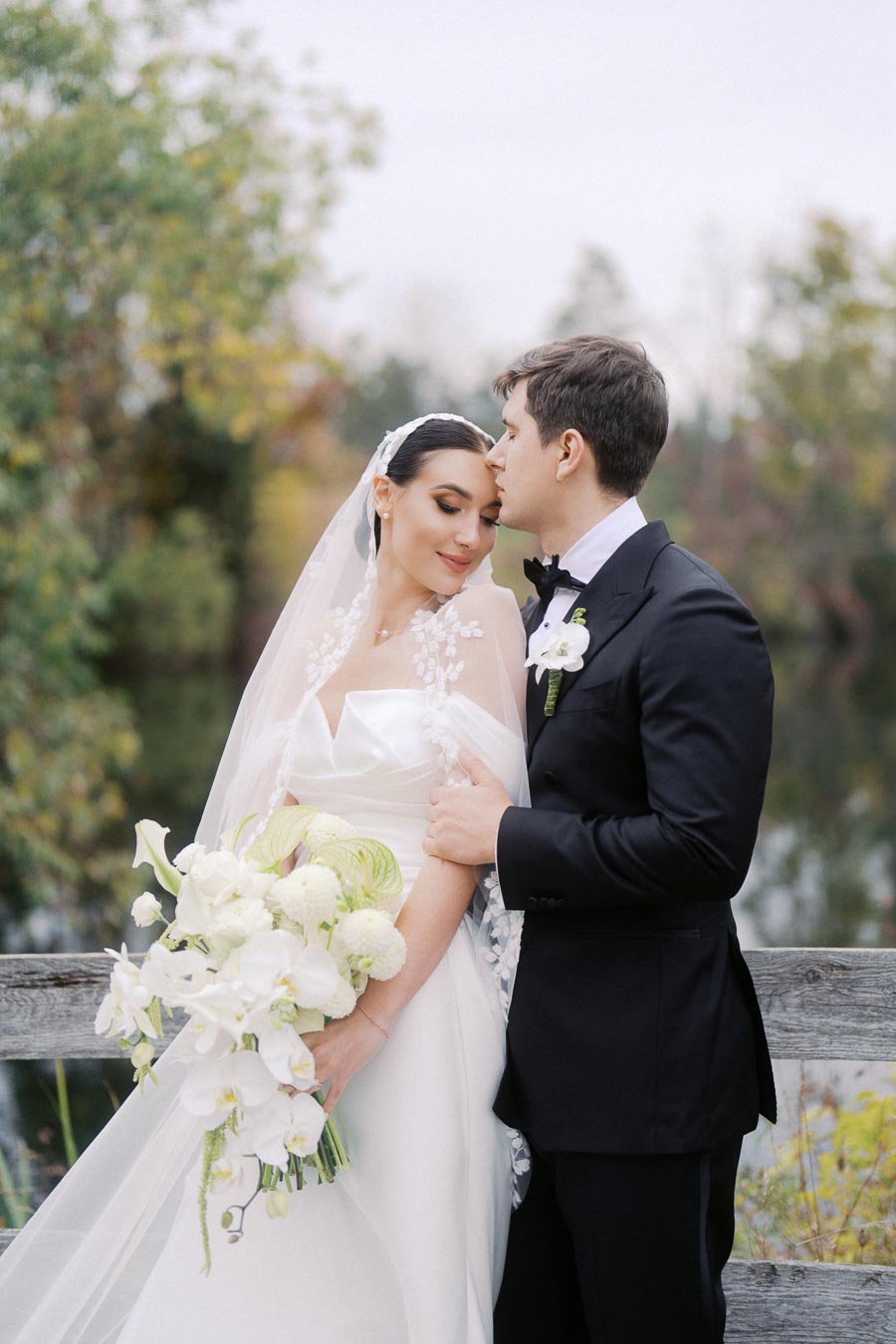 Bride and groom embracing outdoors, bride holding a bouquet of white flowers, wearing a white gown and veil, groom in a black tuxedo, with a blurred natural background.
