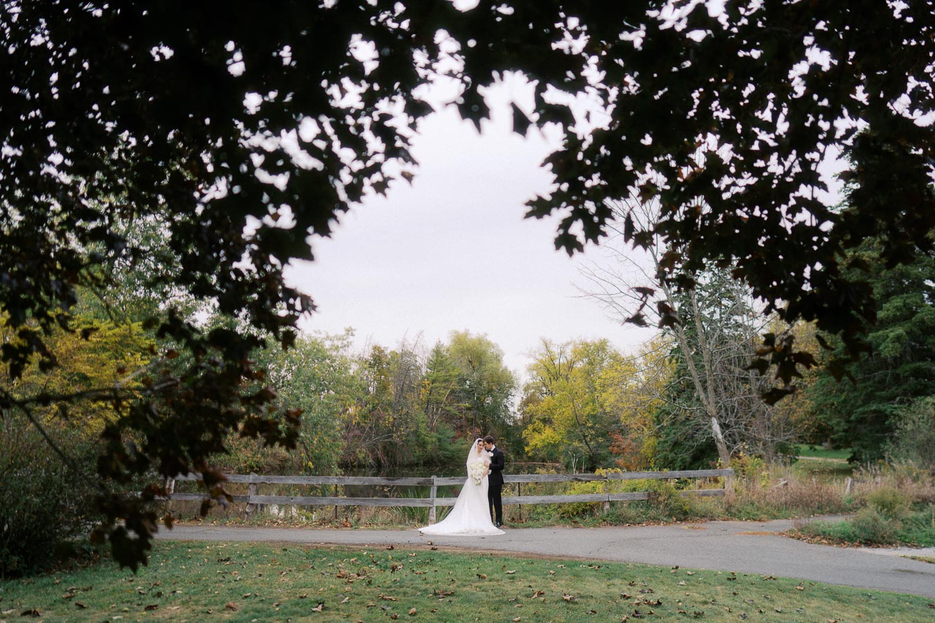 Bride and groom embracing in a picturesque park scene surrounded by lush green trees and a wooden fence, with a natural leaf canopy framing the couple.