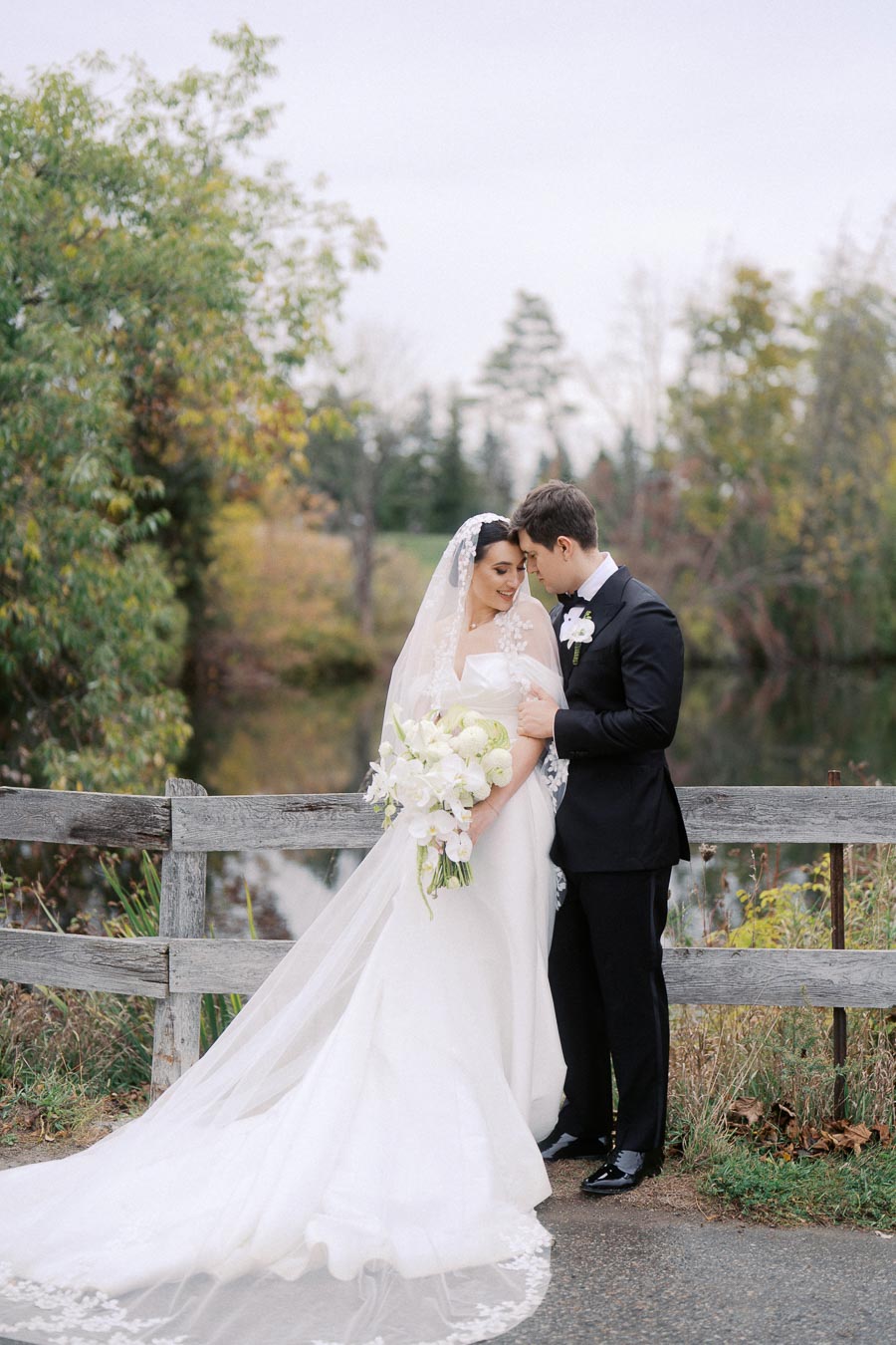 Bride and groom standing by a rustic wooden fence, dressed in elegant wedding attire, with a scenic background of lush greenery and a tranquil pond.