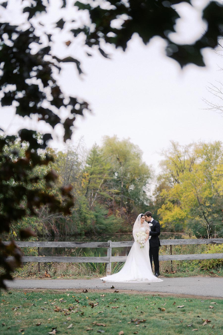 Bride and groom embracing in a serene park setting with lush greenery and a wooden fence, capturing a romantic wedding moment outdoors.