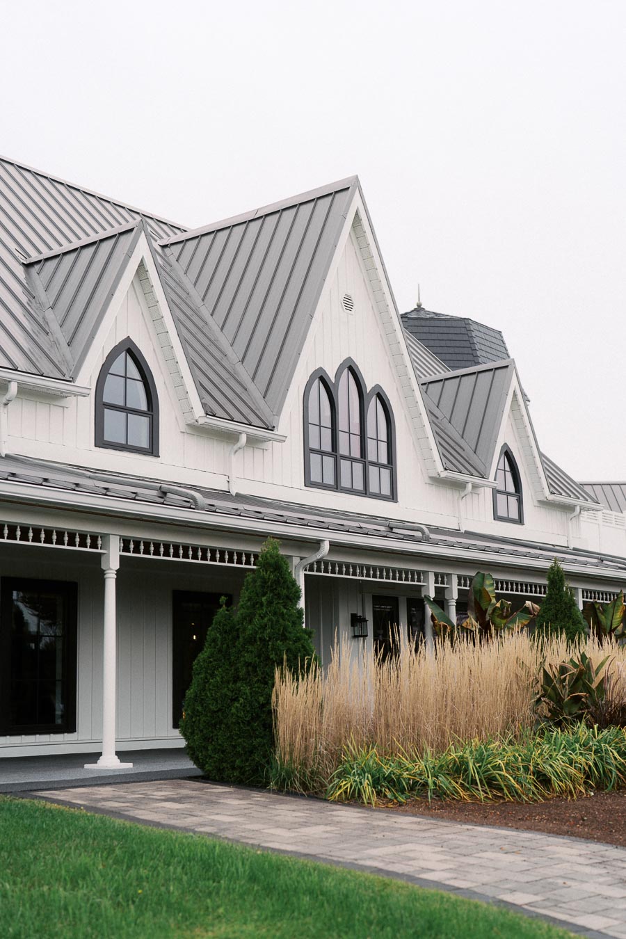 Elegant modern farmhouse with white exterior and gray metal roof, featuring arched windows and lush landscaping with tall grass and shrubs.