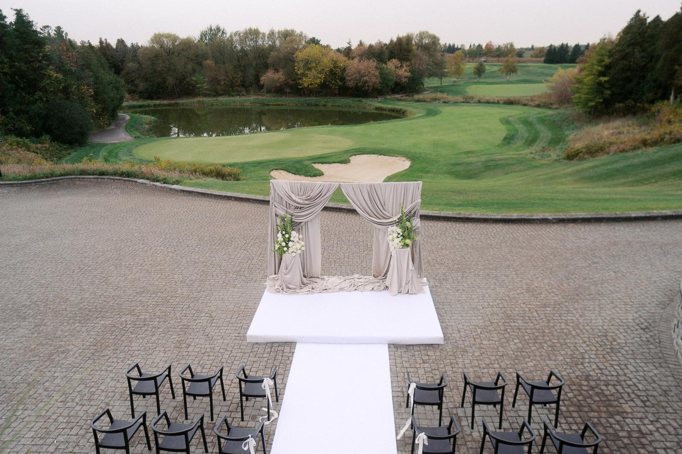 Outdoor wedding ceremony setup with a beautifully draped arch and floral arrangements, overlooking a scenic golf course and pond, with rows of black chairs on a cobblestone platform.