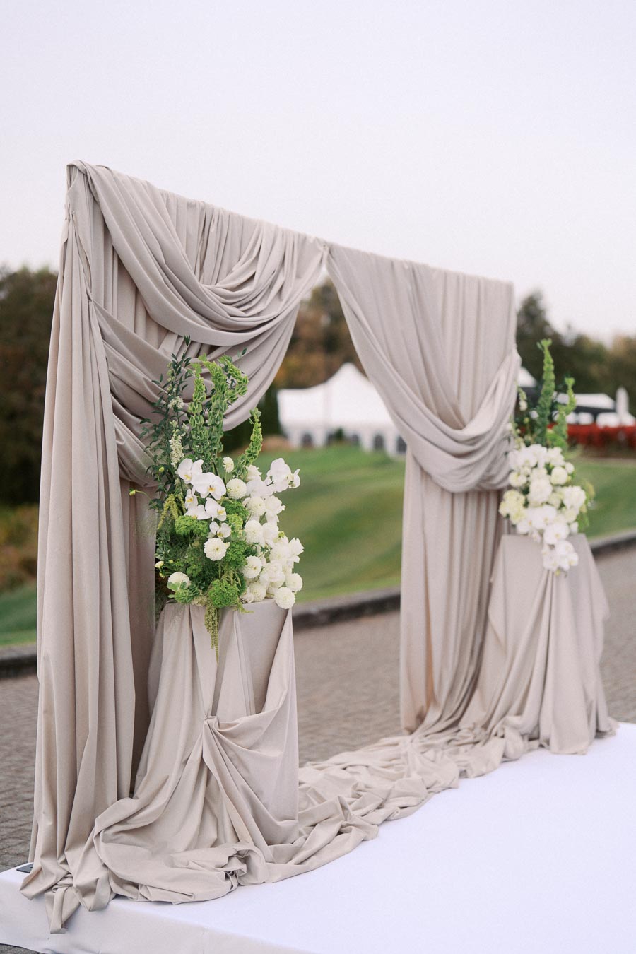 Elegant outdoor wedding arch with flowing beige drapery and white floral arrangements, set against a scenic landscape backdrop.