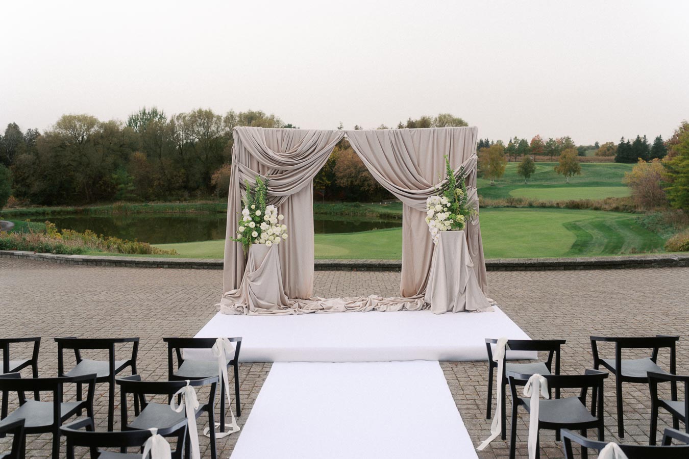 Outdoor wedding ceremony setup featuring a draped arch adorned with white flowers, overlooking a serene landscape with lush greenery and a pond in the background. Black chairs are arranged along a white aisle, creating an elegant and romantic setting.