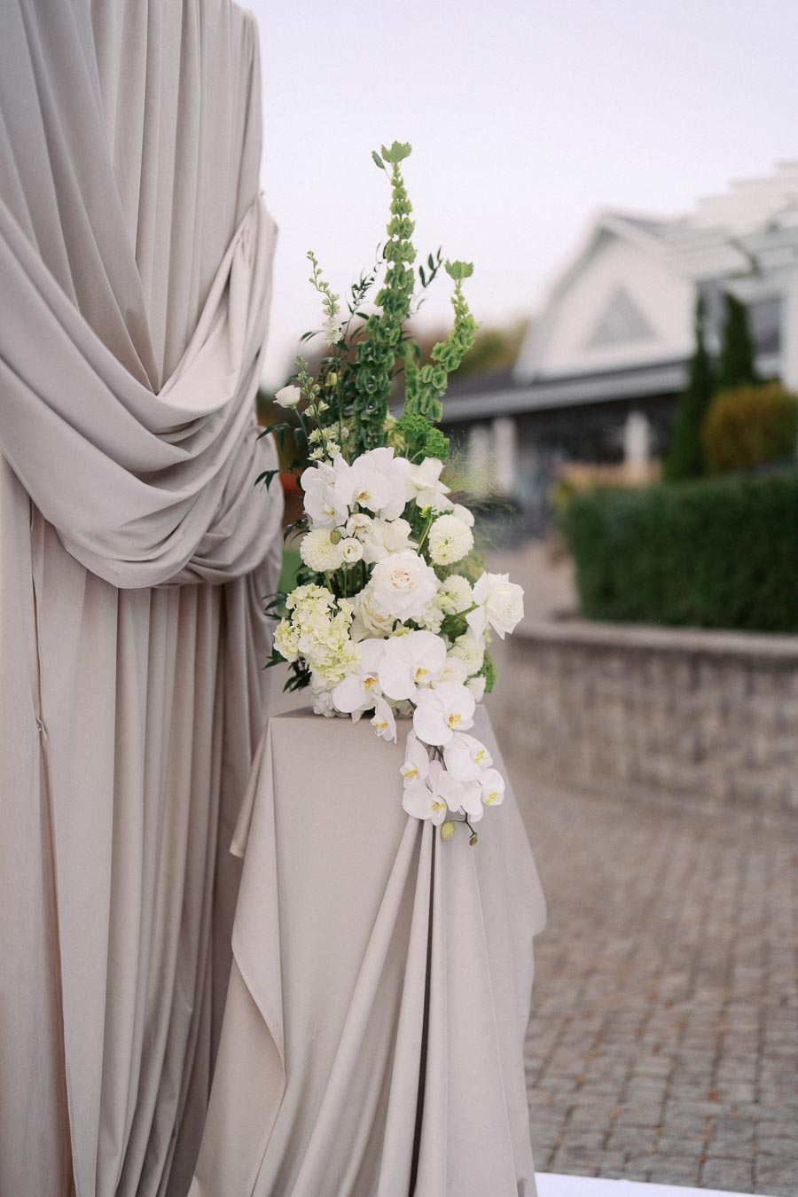 Elegant floral arrangement with white orchids and greenery on a draped pedestal, set against a neutral backdrop, ideal for weddings or events.