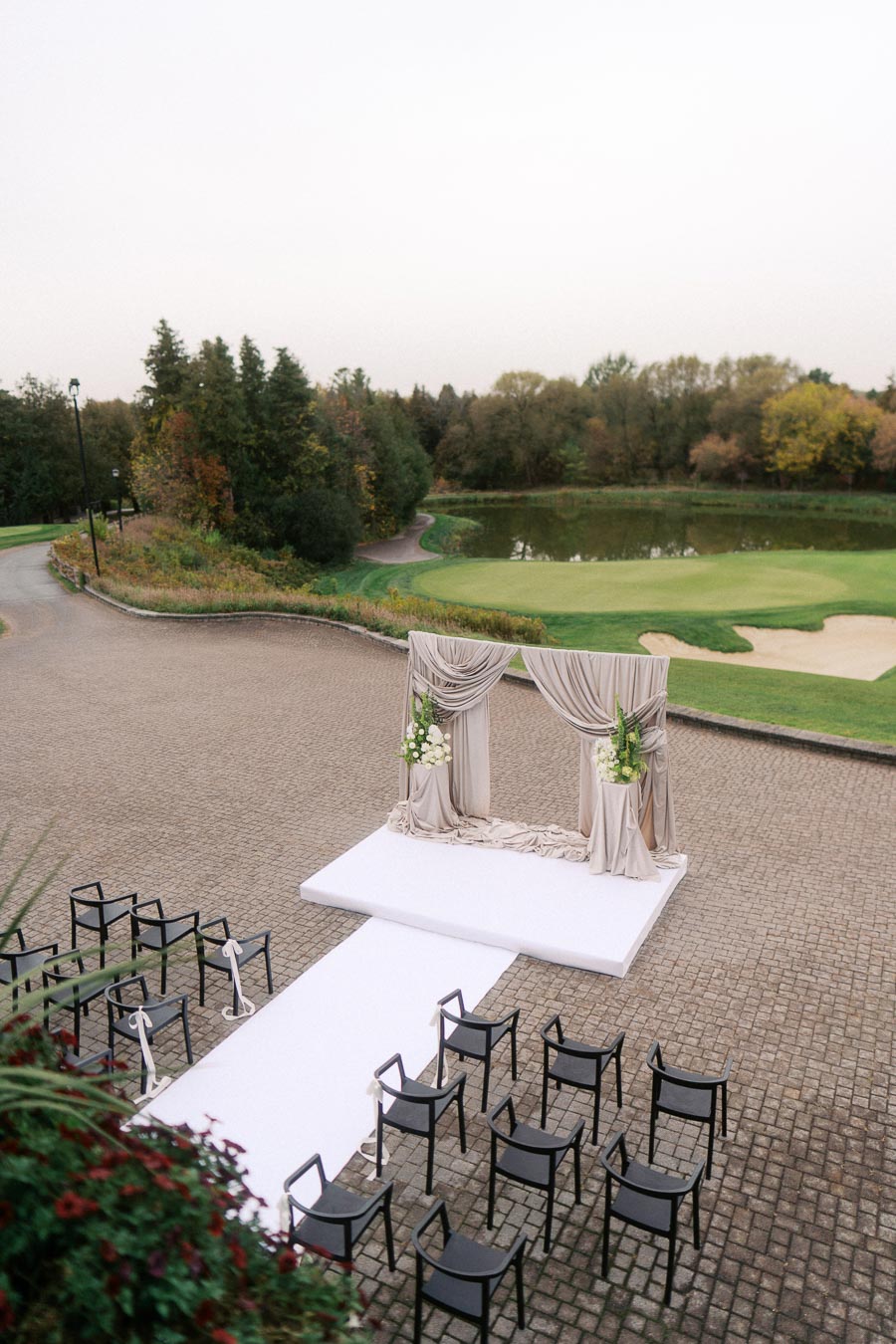 Outdoor wedding ceremony setup on a stone patio, featuring a white aisle runner leading to a draped archway with floral arrangements, surrounded by black chairs, set against a backdrop of lush greenery and a pond.