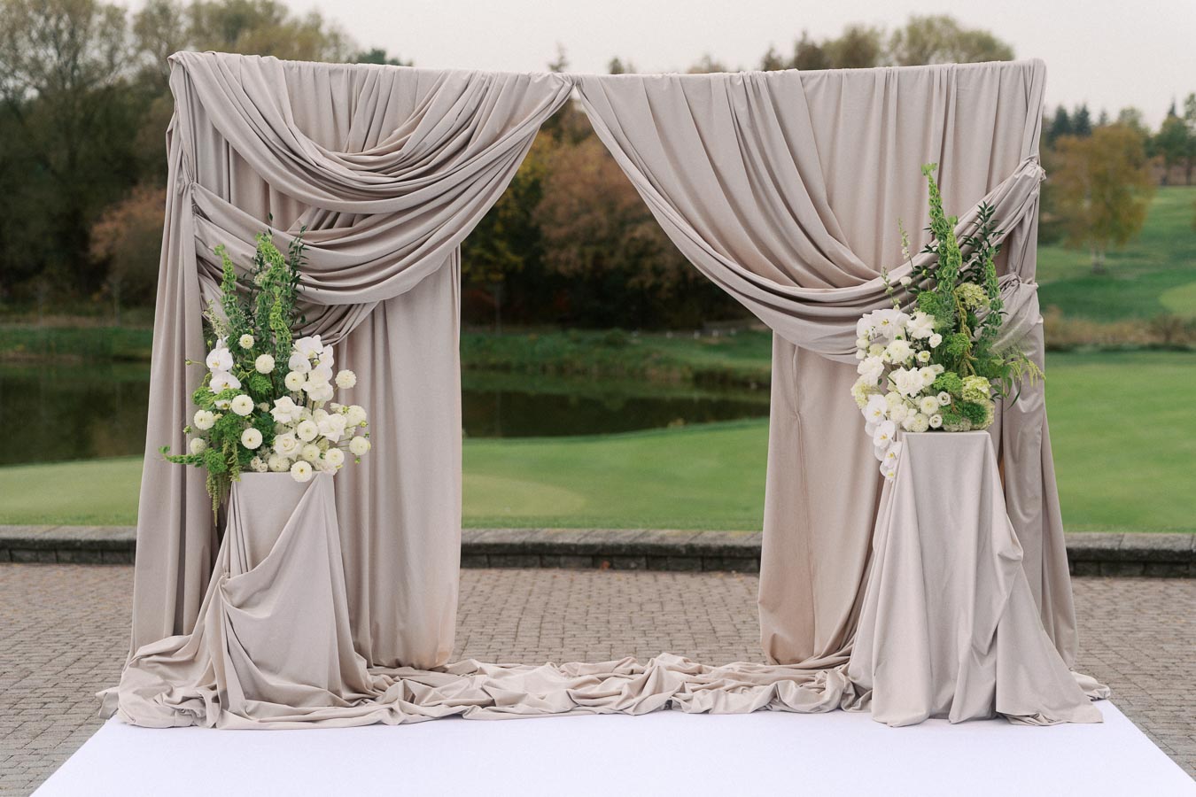Elegant wedding arch with flowing beige drapery and white floral arrangements set against a scenic outdoor backdrop featuring lush greenery and a serene pond.