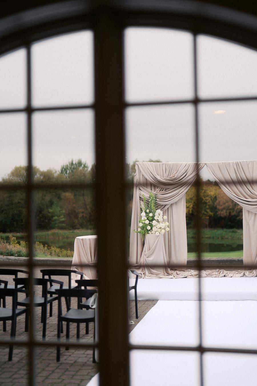 View through a window of an outdoor wedding ceremony setup with draped arches and arranged flowers, featuring elegant black chairs on a patio, surrounded by lush greenery.
