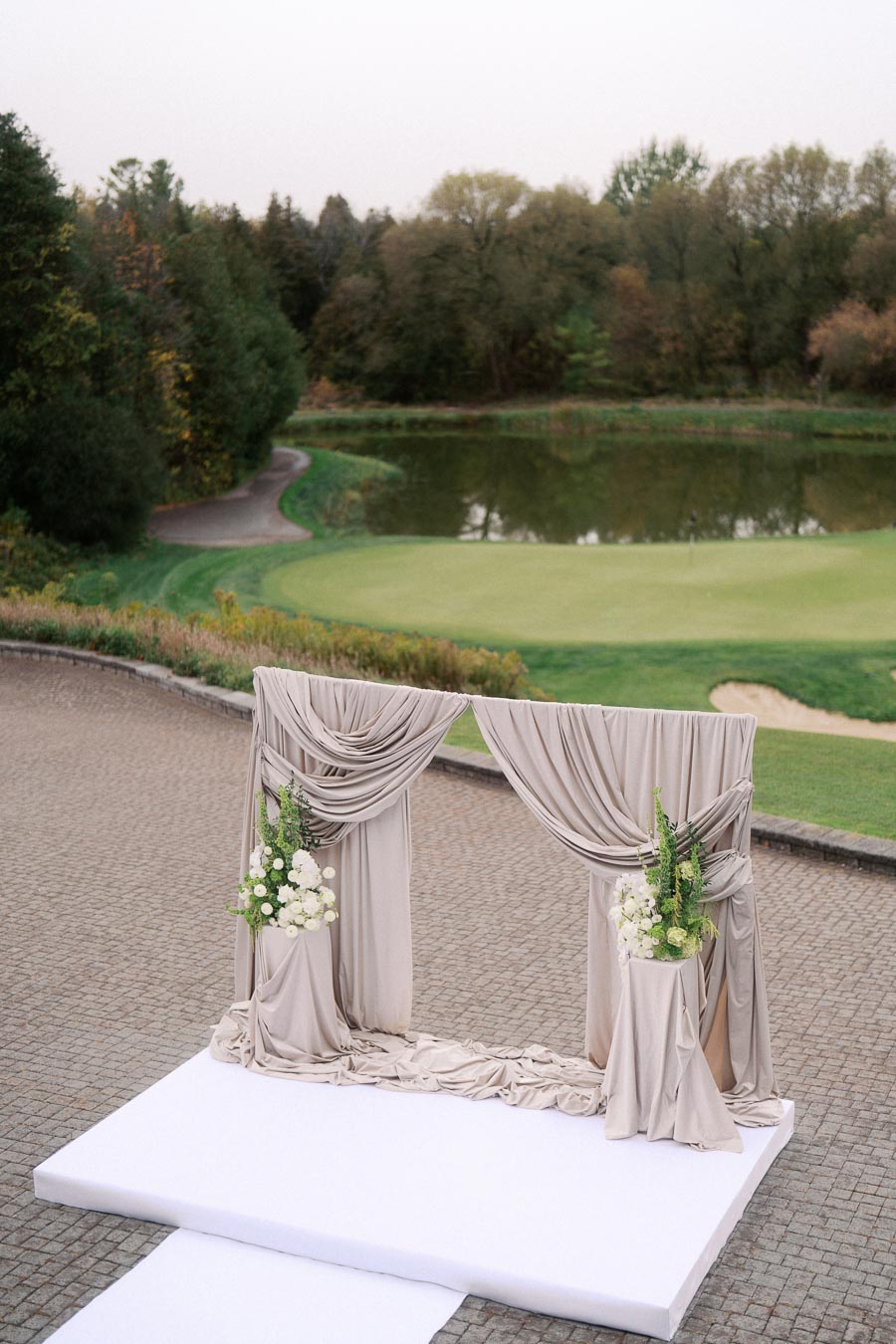 Outdoor wedding altar decorated with elegant beige drapes and floral arrangements, set against a serene golf course landscape with lush greenery and a tranquil pond in the background.