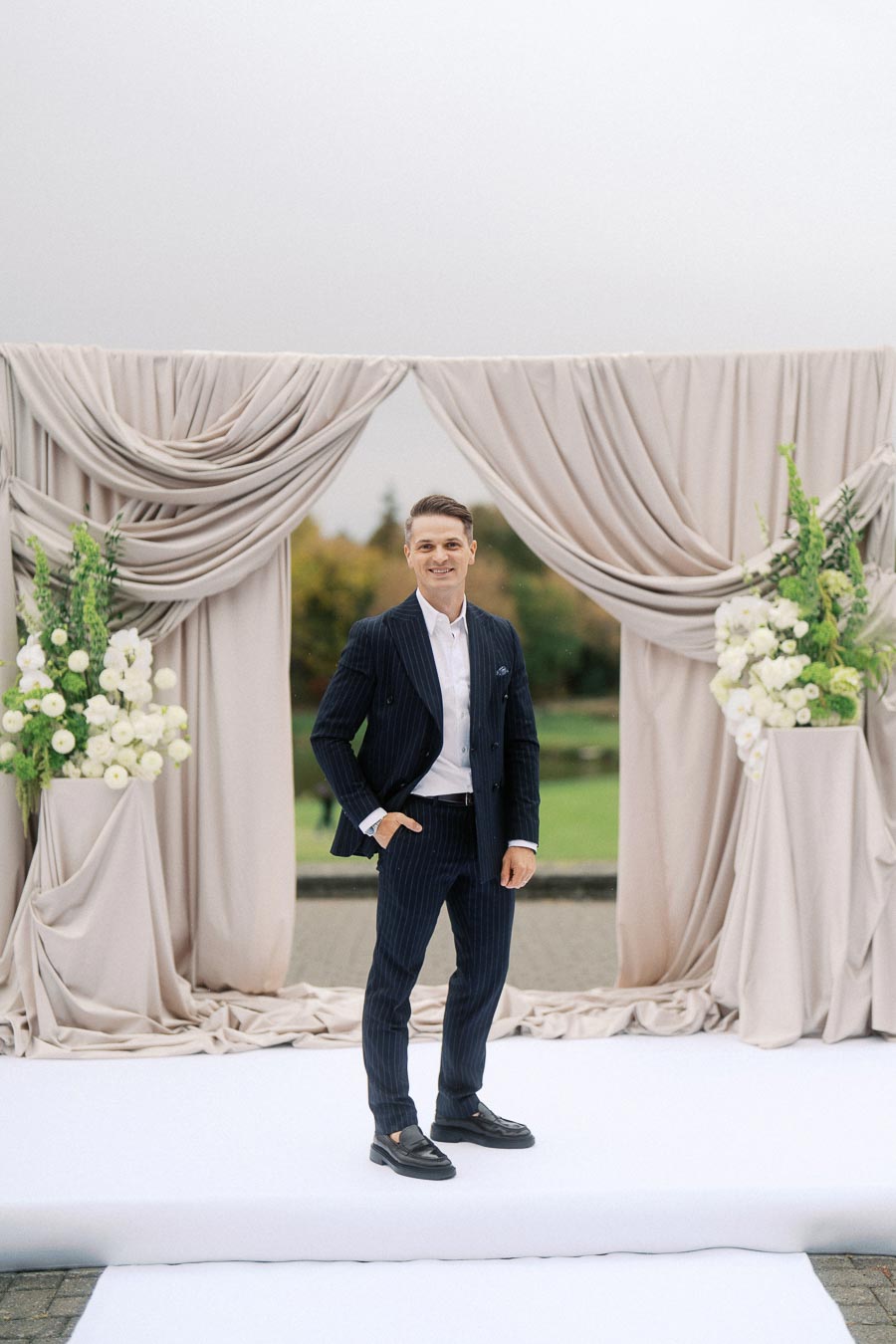 Elegant man in a navy pinstripe suit posing in front of a stylish outdoor wedding backdrop adorned with white and green floral arrangements.