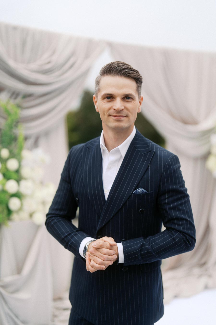 A well-dressed man in a navy pinstripe suit, standing confidently in front of an elegant draped backdrop, which includes greenery and floral arrangements, and smiling at the camera.