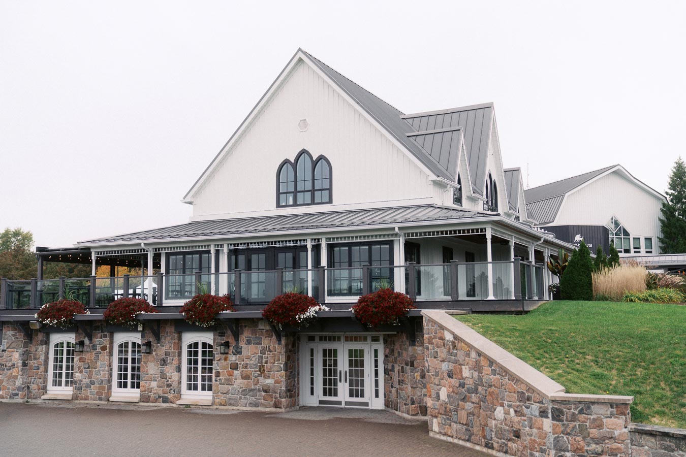 Charming rustic-style venue with stone facade and flower arrangements, featuring large arched windows and a wraparound porch, set against a lush green lawn. Ideal for weddings or events.