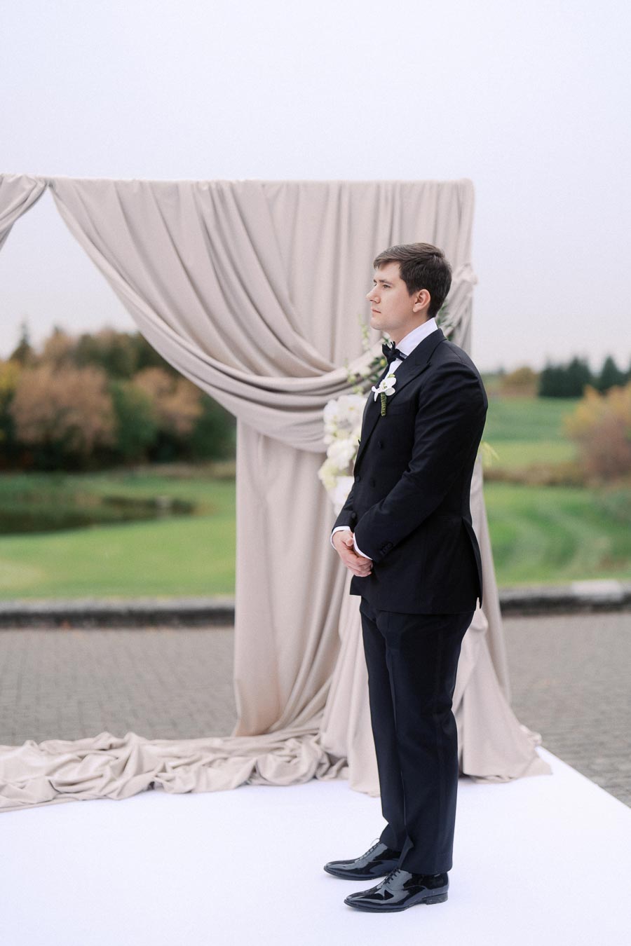Young man in a formal black suit standing outdoors, in front of an elegant draped fabric backdrop, with lush greenery in the background on a cloudy day.