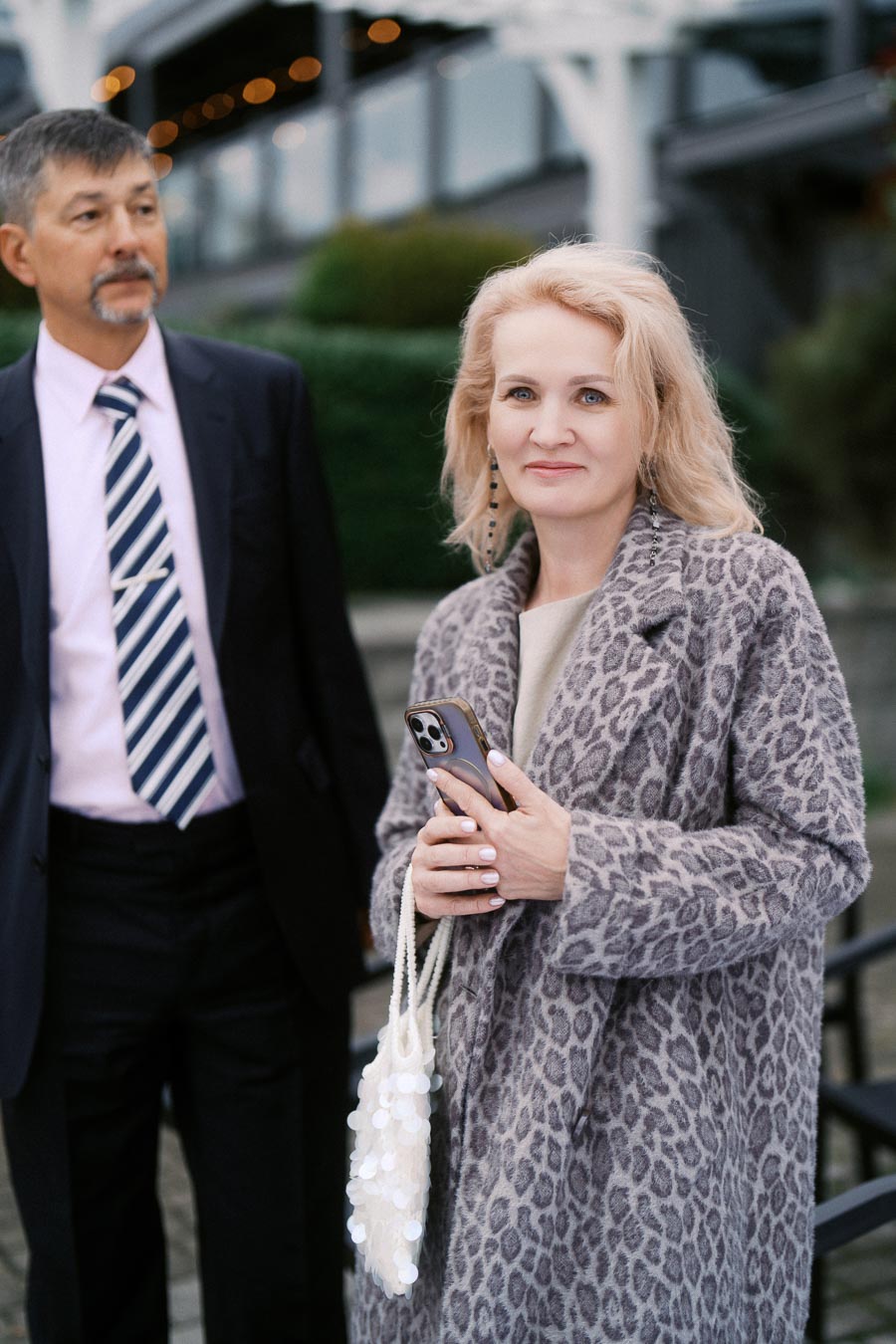 A woman in a leopard print coat holding a smartphone, standing outdoors next to a man in a suit with a striped tie, with a blurred urban background.