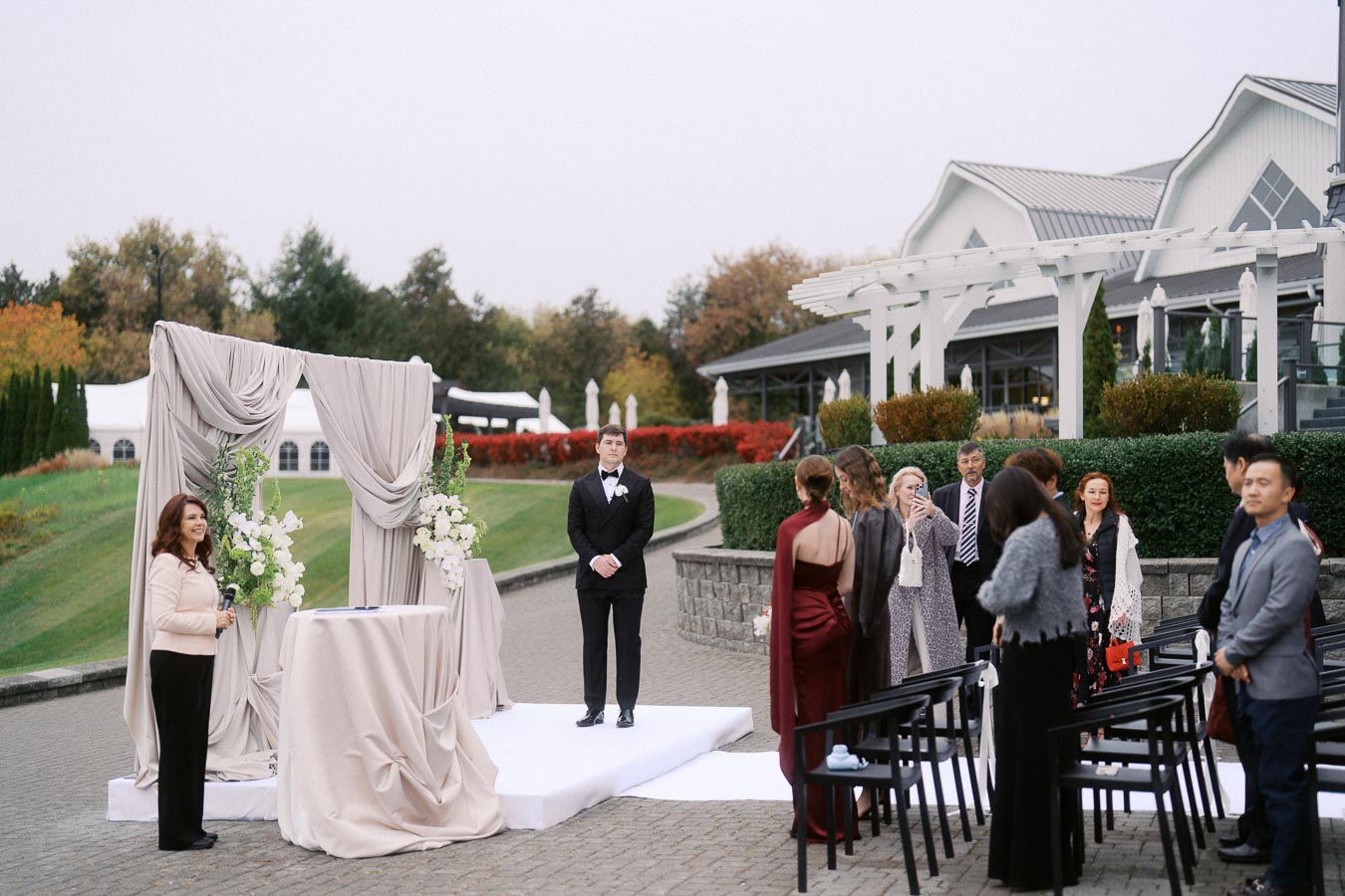 Outdoor wedding ceremony setup with a groom in a tuxedo standing at the altar, decorated with draped fabric and floral arrangements. Guests are gathered, dressed in formal attire, against the backdrop of a picturesque venue with green lawns and autumn foliage.