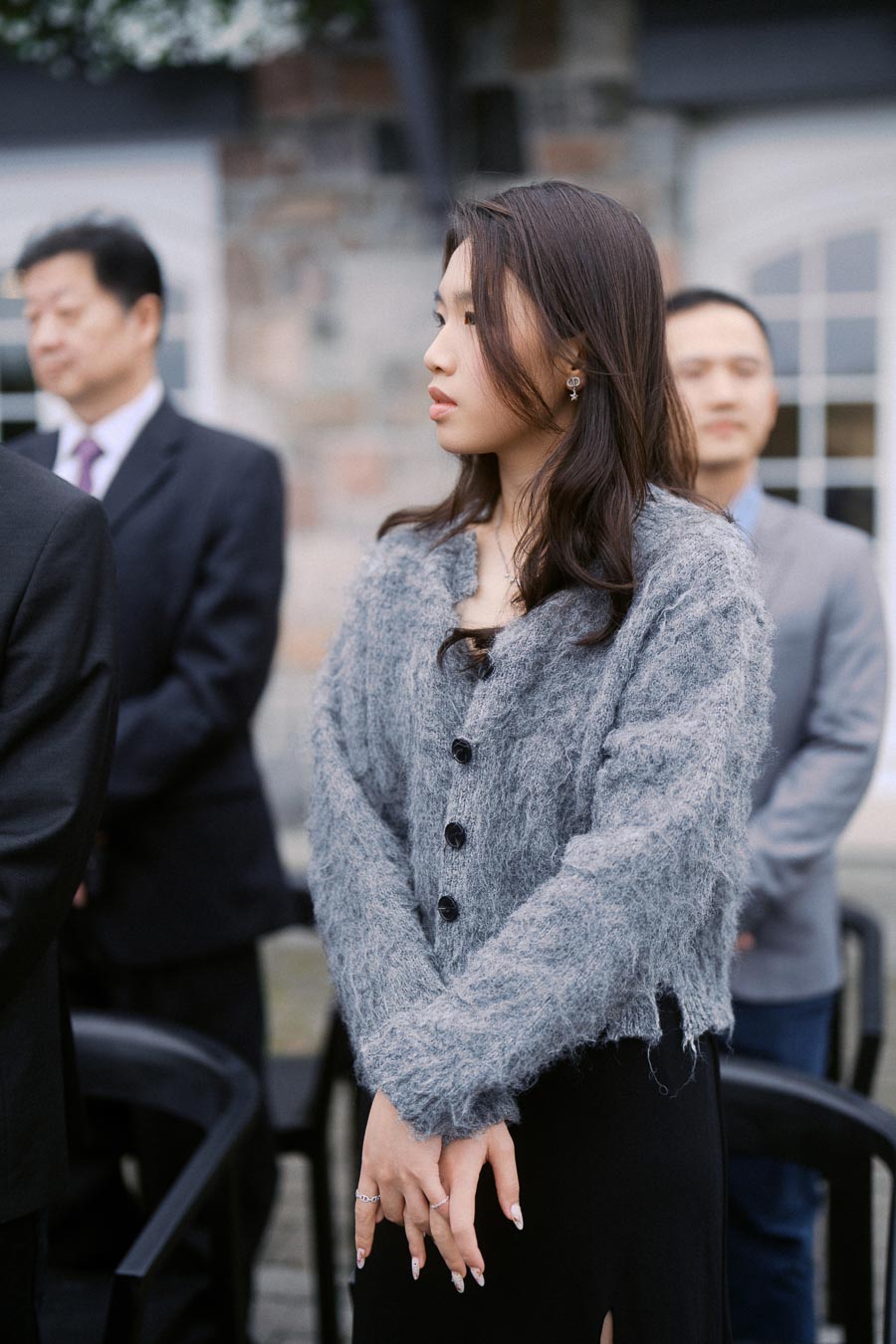 Woman in a gray cardigan attends outdoor formal event, standing attentively in a group.