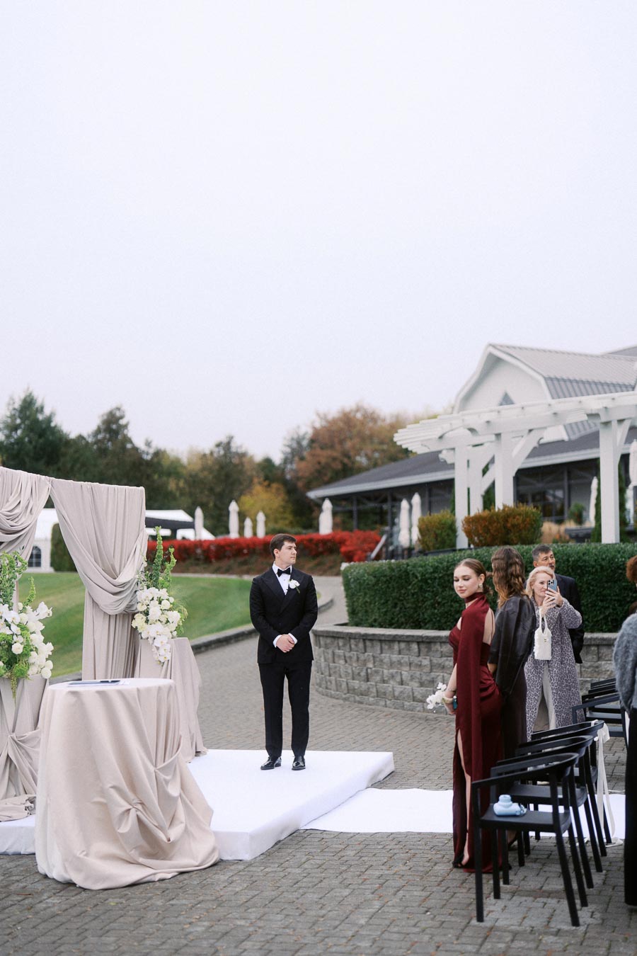 Outdoor wedding ceremony setup with a groom in a tuxedo standing on a decorated platform. Bridesmaids in elegant dresses and guests are gathered around, with a backdrop of landscaped greenery and a white pergola. Relaxed and elegant atmosphere.