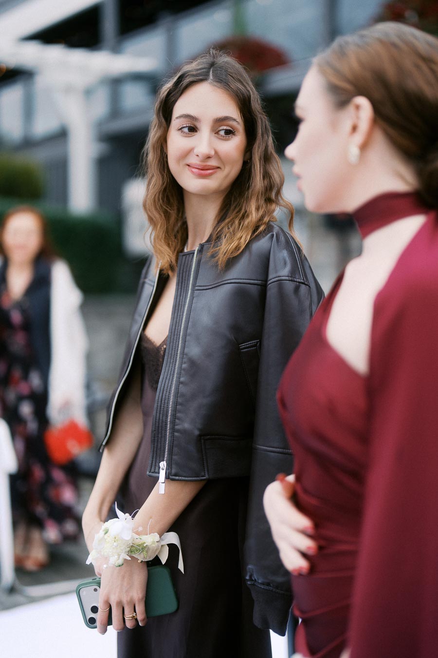 Two women in elegant attire sharing a conversation at an outdoor event, one wearing a black jacket and holding a smartphone with a floral corsage on her wrist, and the other in a burgundy dress.