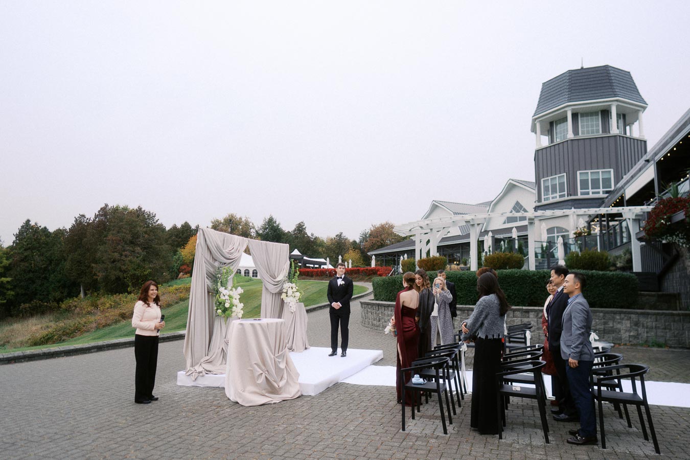 Outdoor wedding ceremony set up with elegant draped archway and floral arrangements. Guests stand near a cobblestone path outside a modern building in a scenic location.