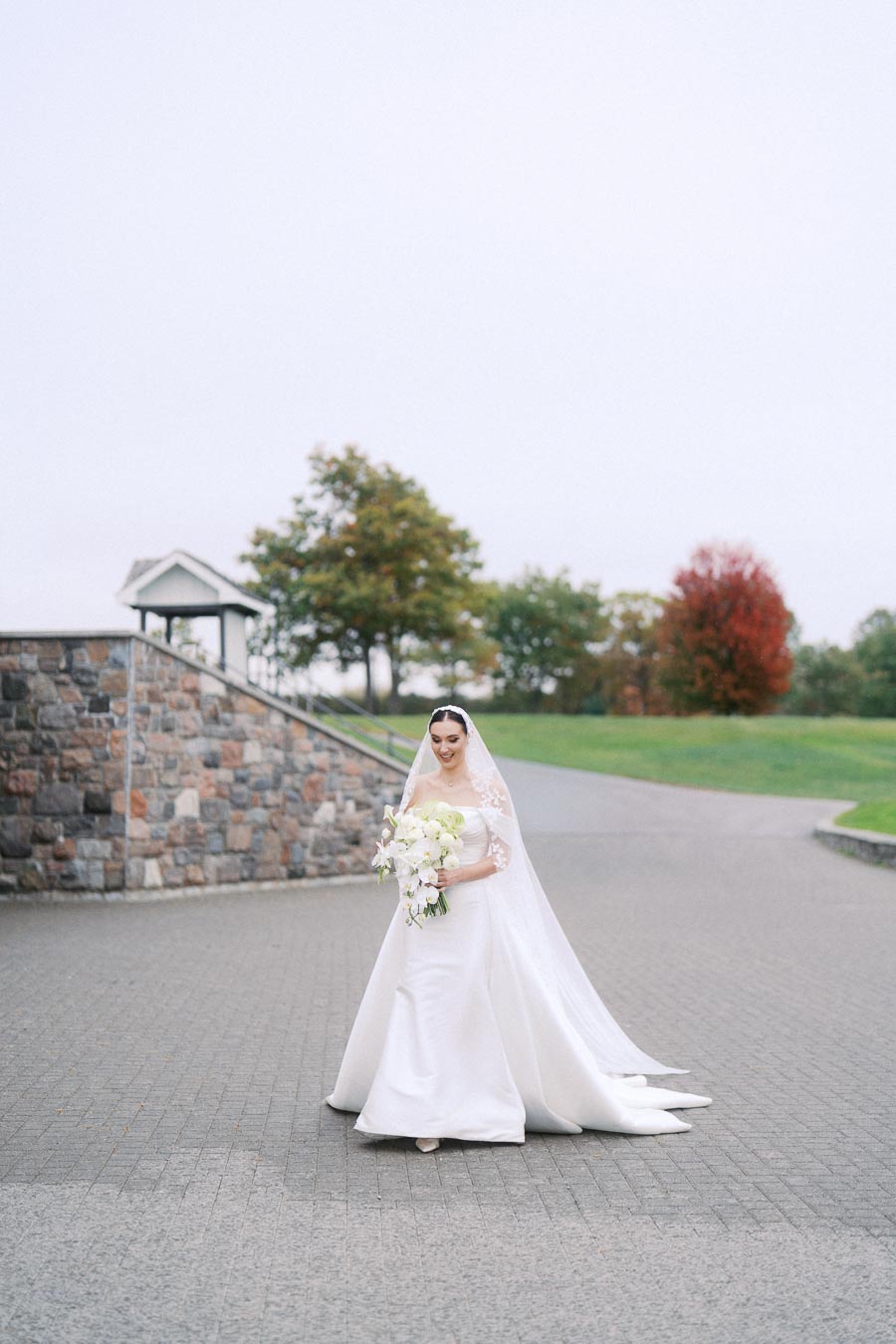 Bride in elegant white wedding dress and veil holding a bouquet of white flowers, standing on a paved path with greenery and a stone wall in the background, showcasing an outdoor wedding setting.