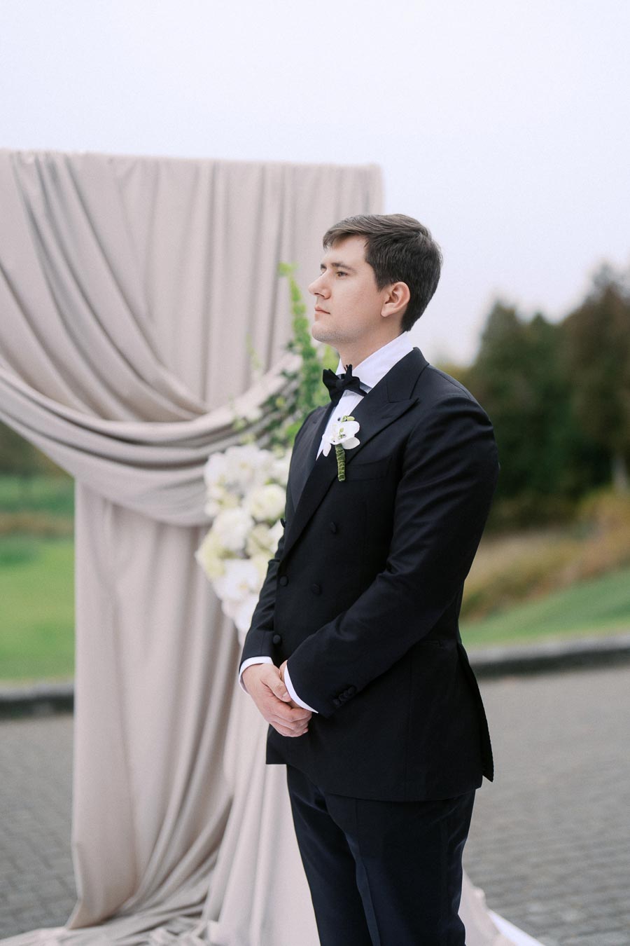Man in elegant black tuxedo standing in front of a draped wedding arch with floral decorations outdoors.
