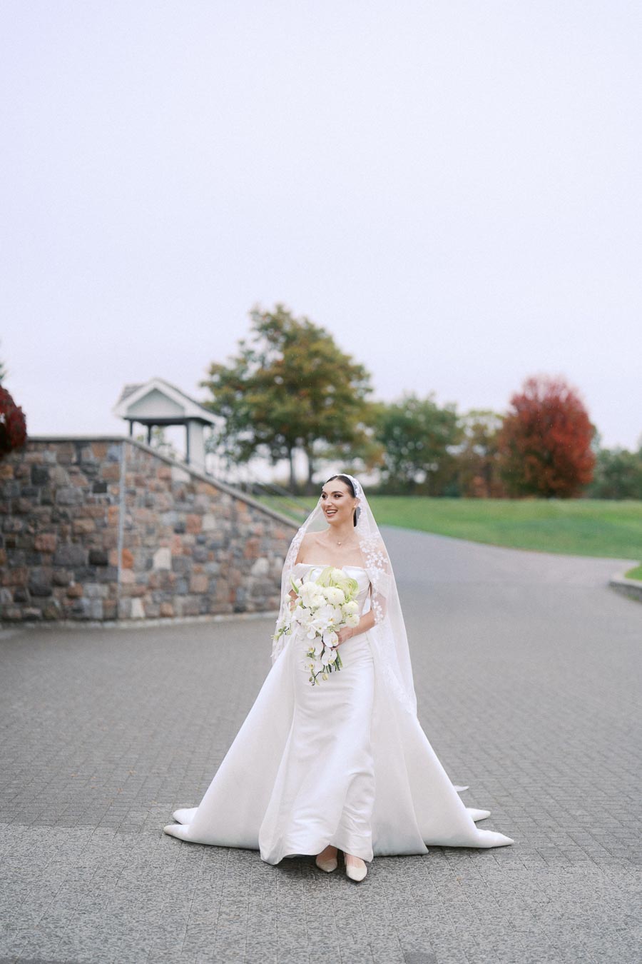 Bride in an elegant white wedding dress with lace veil holding a bouquet, walking on a stone pathway in a scenic outdoor setting.