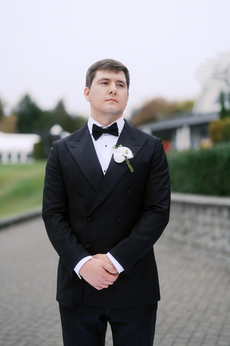 A man in a tuxedo with a white boutonniere, standing outdoors on a paved path, looking confidently to the side.