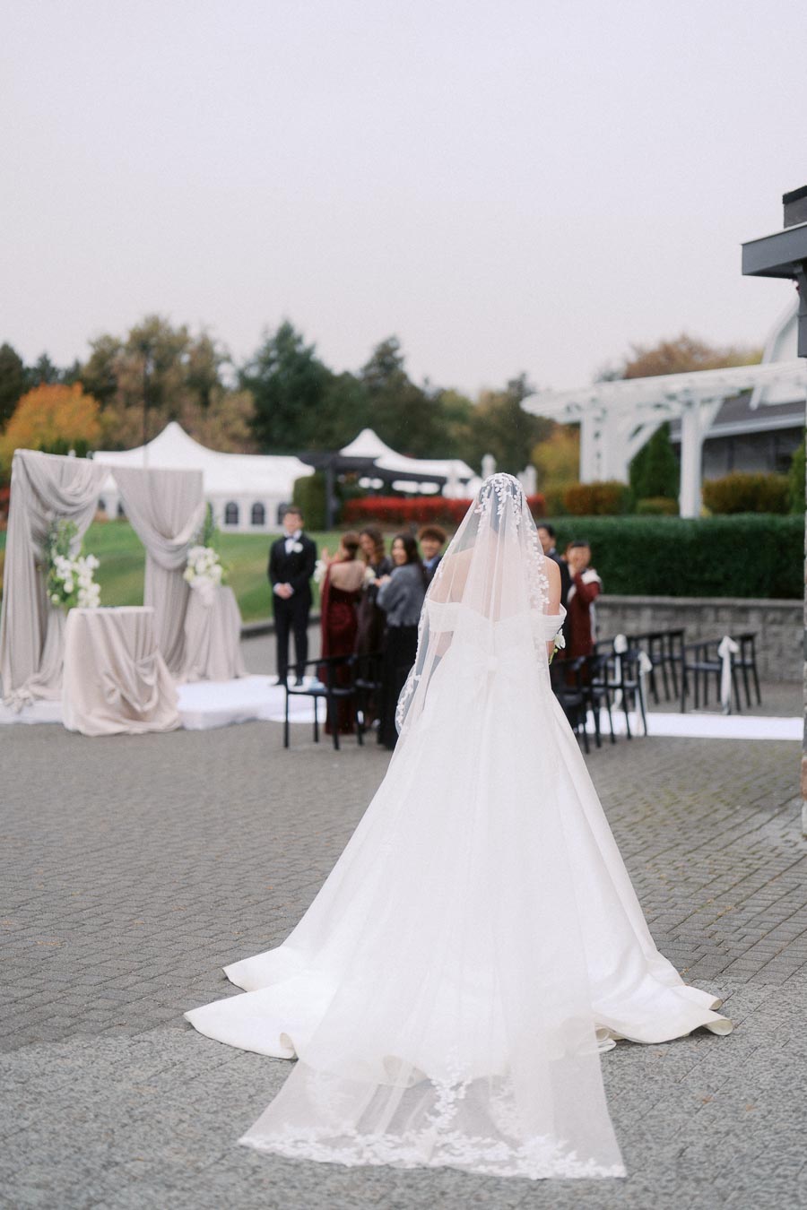 A bride in a white gown and veil walks towards an outdoor wedding ceremony setup, featuring a draped altar and guests seated, with a garden and pavilion in the background.