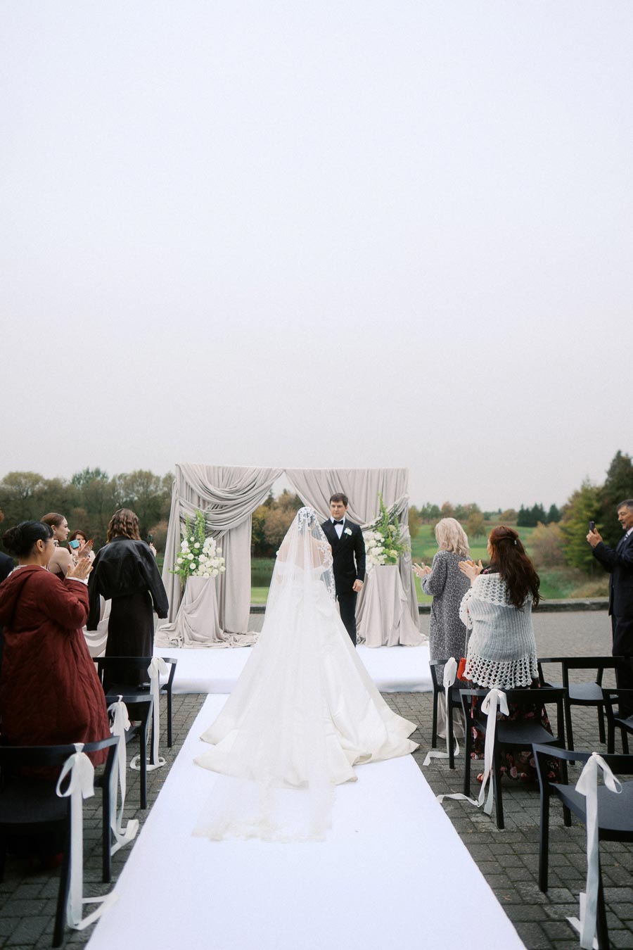 Bride walking down an outdoor aisle towards a groom in a tuxedo, with guests applauding by a beautifully decorated wedding arch.