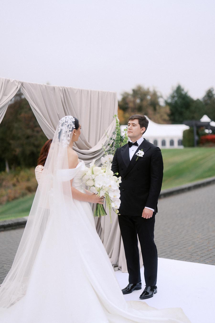 Bride and groom standing at an outdoor wedding ceremony under a draped archway, with the bride holding a bouquet of white flowers and the groom in a black tuxedo, set against a backdrop of greenery and a white tent.