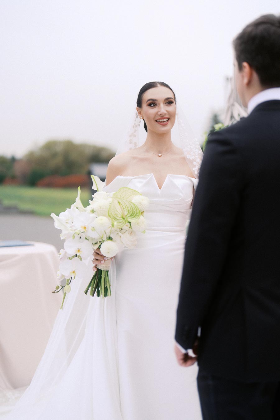 Bride in elegant white wedding dress holding a bouquet of white flowers and smiling at groom outdoors.