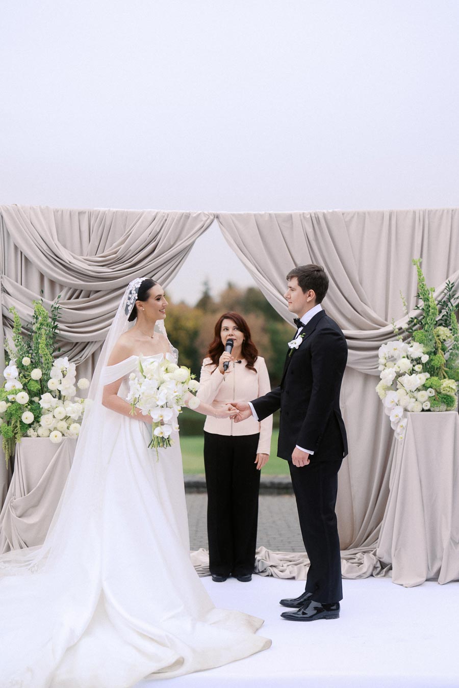 Bride and groom holding hands during an outdoor wedding ceremony, with an officiant speaking, surrounded by elegant white floral arrangements and draped decor.