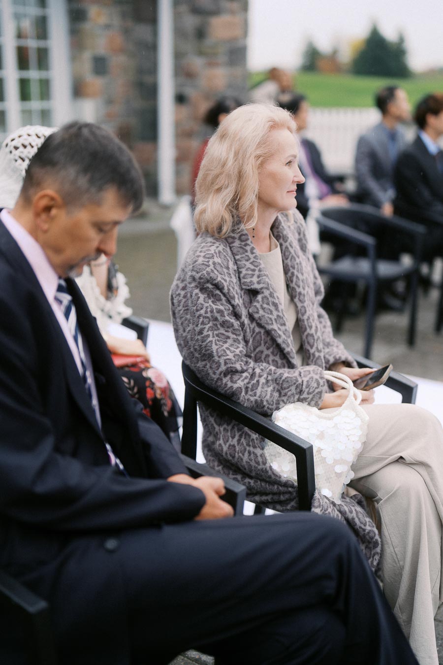 An elegantly dressed woman in a leopard print coat sits attentively in an outdoor event, holding a white bag, surrounded by other seated attendees.