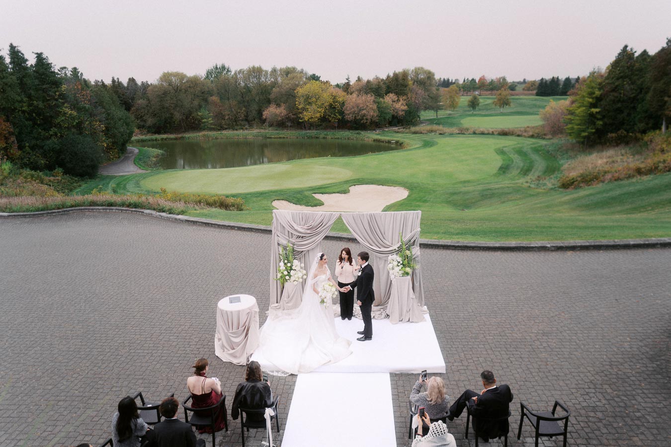Outdoor wedding ceremony at a golf course featuring a couple exchanging vows under a draped archway with a scenic view of lush green fairways and a tranquil pond, surrounded by a small gathering of guests.