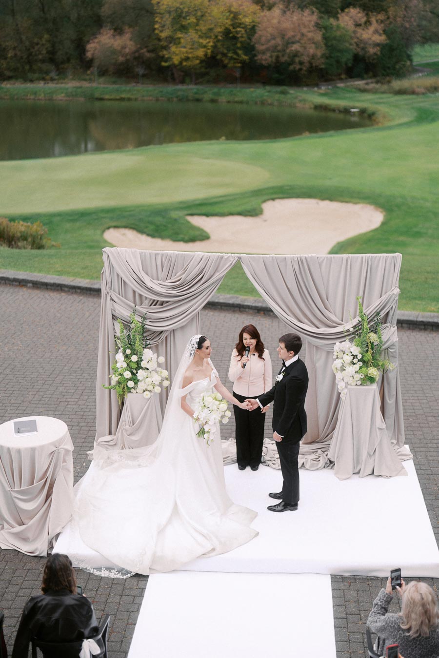 Elegant outdoor wedding ceremony with a bride and groom standing under a draped fabric arch, exchanging vows by a scenic golf course, surrounded by white floral arrangements, with guests capturing the moment.