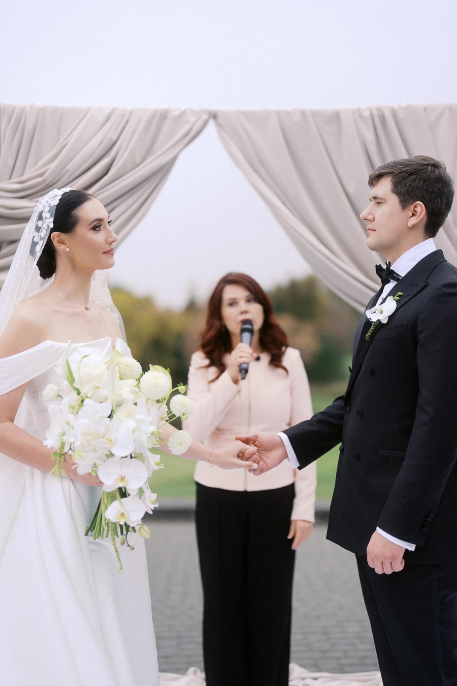 Bride and groom exchanging vows during an outdoor wedding ceremony with an officiant in the background, elegant floral arrangement in the bride's hands.