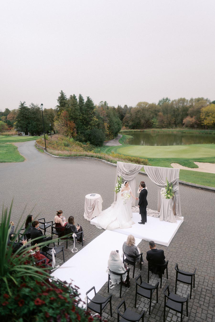 Aerial view of an outdoor wedding ceremony at a serene golf course, featuring a bride and groom exchanging vows under a draped archway, surrounded by a small group of seated guests, with lush greenery and a pond in the background.