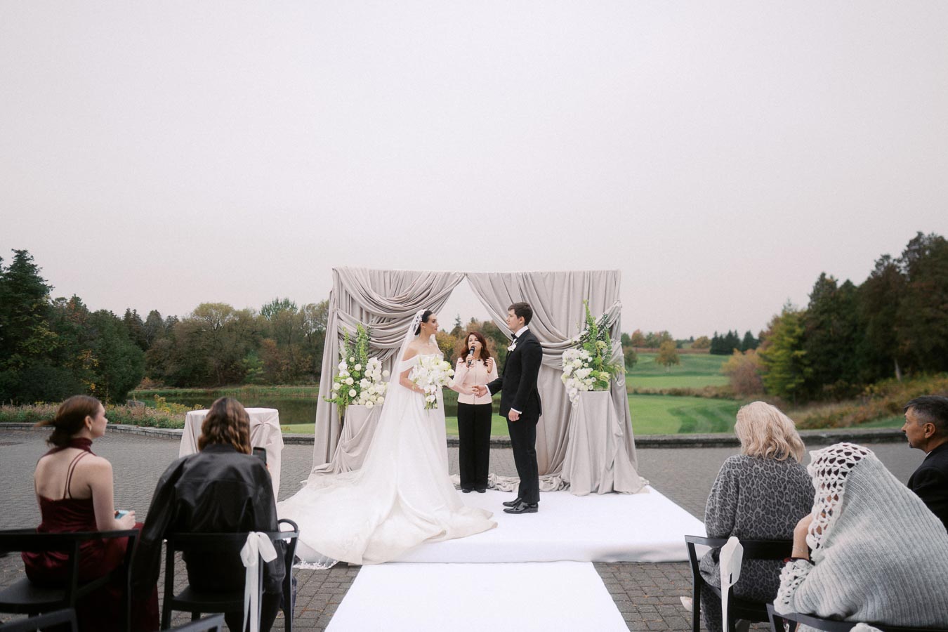 Outdoor wedding ceremony with a bride and groom under a decorated archway, surrounded by lush greenery. Guests are seated in the foreground, witnessing the couple exchange vows in a scenic natural setting.