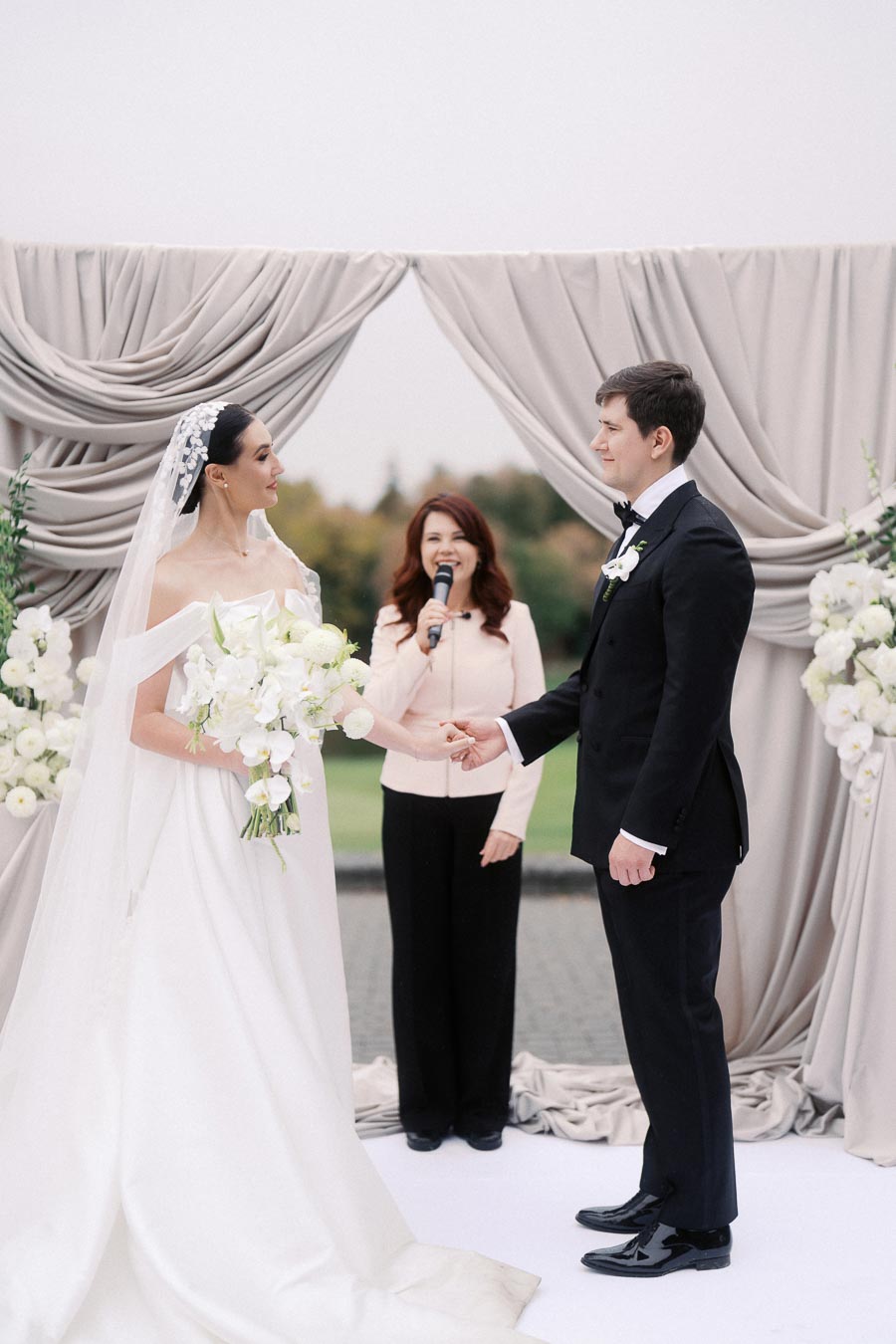 Bride and groom holding hands during outdoor wedding ceremony with officiant, surrounded by elegant floral decorations.