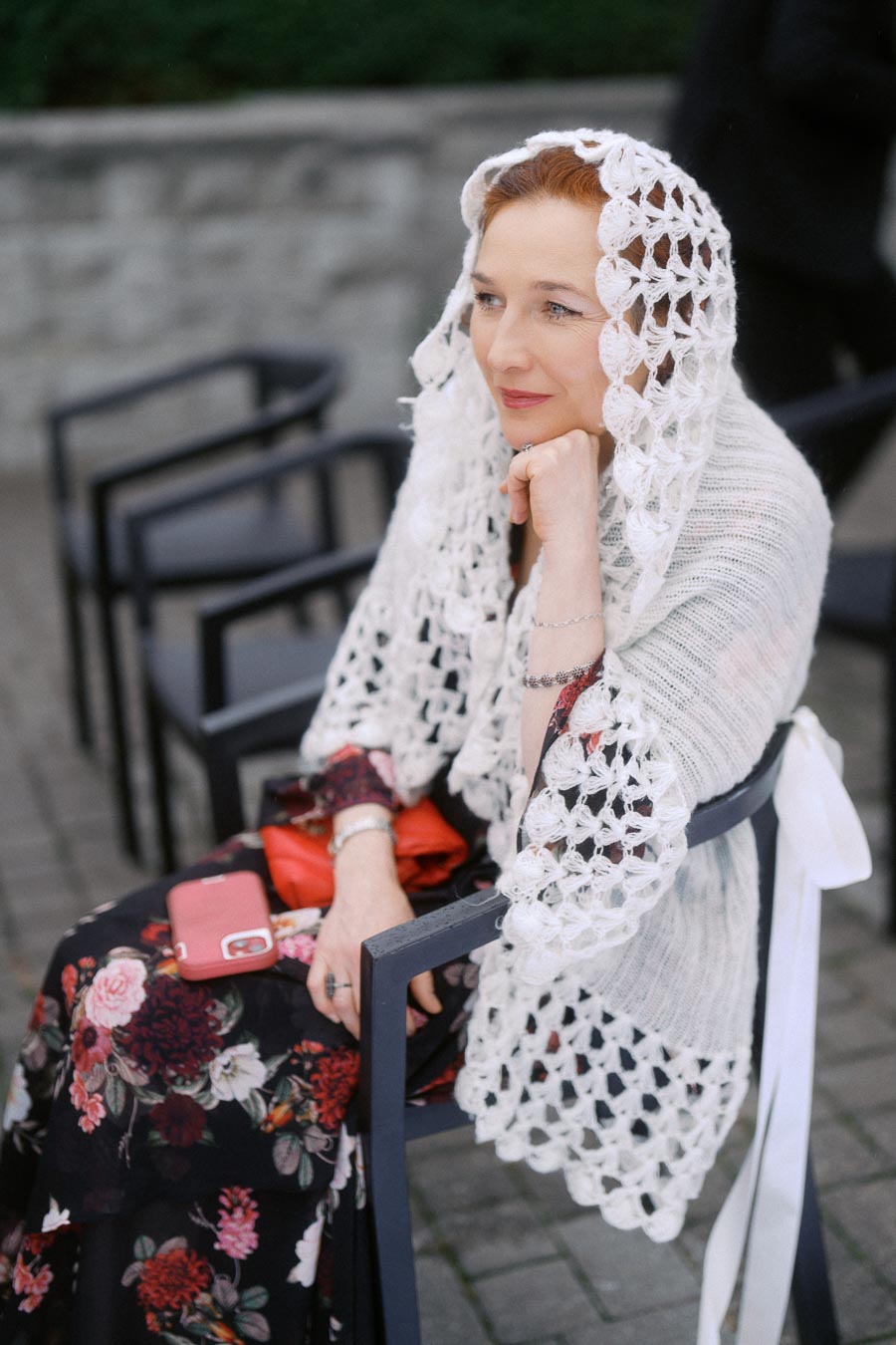 Woman in a floral dress and white crochet shawl sitting outdoors, looking thoughtfully into the distance with a smartphone beside her.